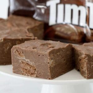 Pieces of chocolate fudge with chunks of Tim Tam biscuits throughout on a white cake stand.