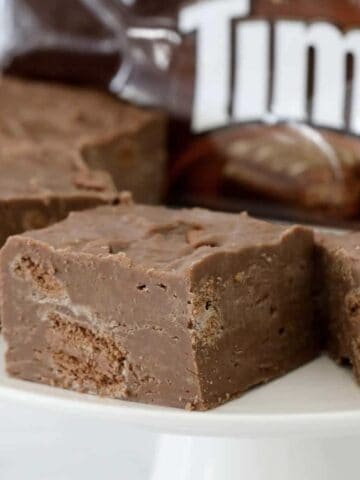 Pieces of chocolate fudge with chunks of Tim Tam biscuits throughout on a white cake stand.