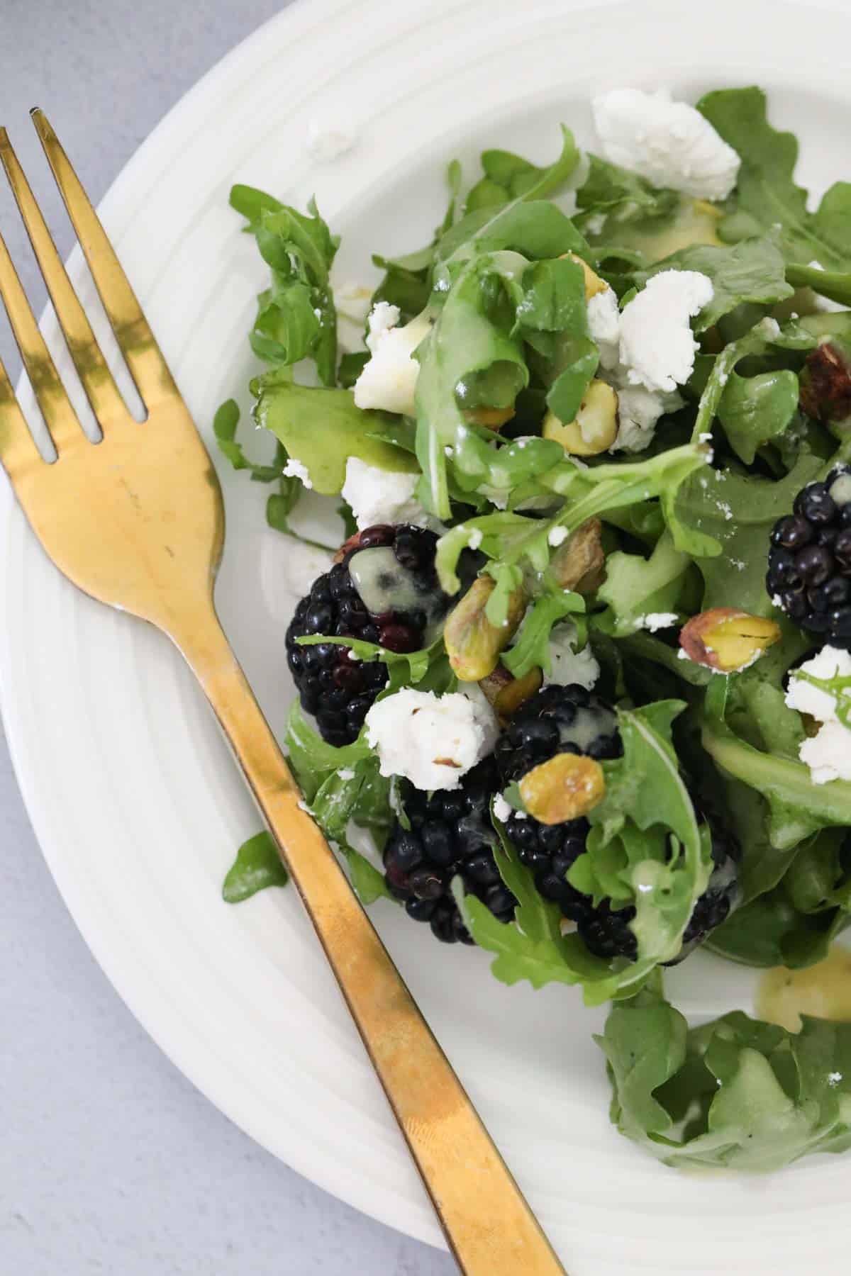 Looking down on a white plate with blackberry, feta, pistachio and rocket salad, with a gold fork on the side.