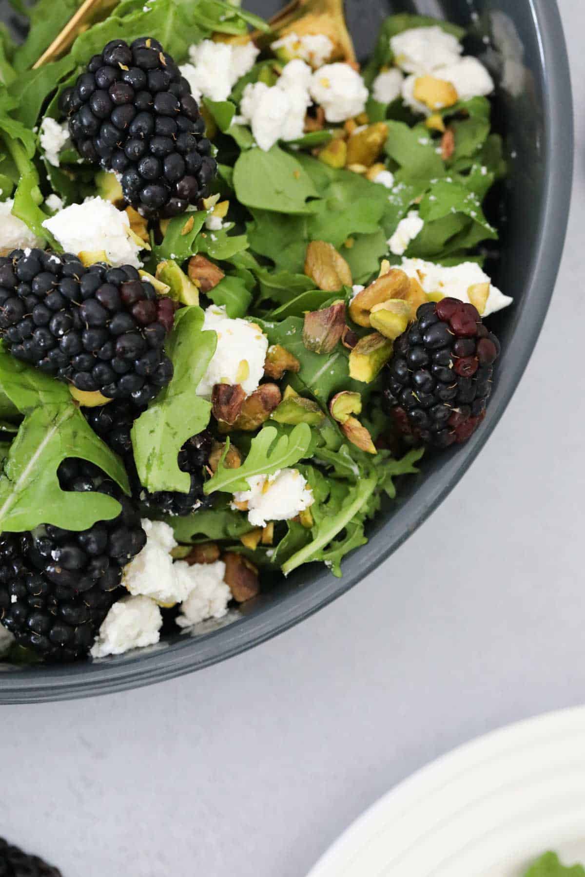 Looking down on a black bowl with crumbled feta and blackberries on green salad leaves.