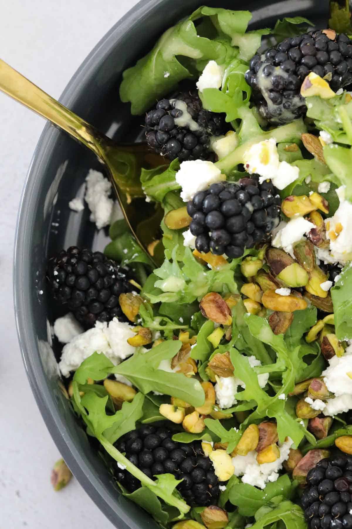 A black salad bowl filled with rocket, blackberries, pistachios and crumbled feta.