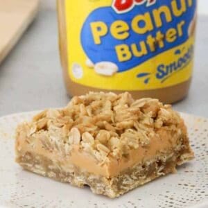 A piece of peanut butter and oat slice in front of a jar of peanut butter.
