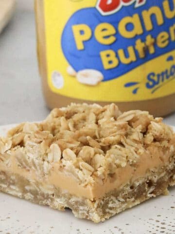A piece of peanut butter and oat slice in front of a jar of peanut butter.