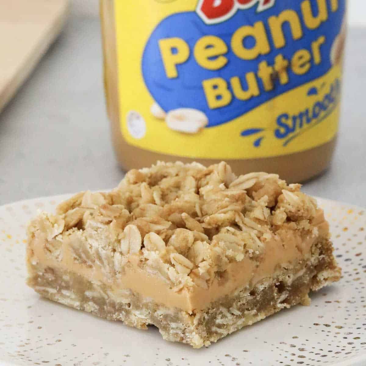 A piece of peanut butter and oat slice in front of a jar of peanut butter.